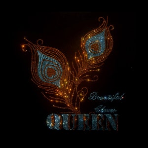 以下が含まれることがあります： 黒のTシャツに、ラインストーンでできた孔雀の羽根2本のデザインが施されています。羽根はオレンジとブルーです。"Beautiful Never"と"QUEEN"の文字もラインストーンでできています。