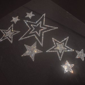 Può includere: Una superficie in tessuto nero con sette motivi a forma di stella realizzati con strass trasparenti e riflettenti. Le stelle variano in dimensioni e sono disposte in modo sparso, ciascuna delineata da un bordo denso di cristalli scintillanti.