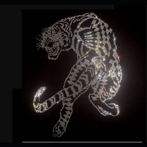 Può includere: Un disegno di tigre composto da numerosi piccoli strass argentati riflettenti su uno sfondo nero solido. La tigre è raffigurata in una postura arcuata e in agguato, con la testa abbassata e la coda curva, che mostra un dettagliato motivo a strisce.