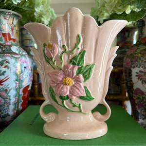 May include: A pale pink ceramic vase with a scalloped rim and a raised relief design featuring a pink flower with a yellow center and green leaves. The vase has two small curved handles at the base and a smooth, glossy finish on its surface.