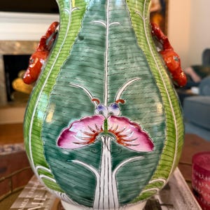 May include: A ceramic vase featuring a green leaf pattern with a central pink and purple butterfly motif. The vase has a white base and two orange, sculpted animal-shaped handles on the sides. The surface is glossy with visible painted details.