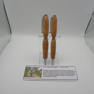 May include: Two Extra Fancy Eucalyptus wood pens with silver-colored accents. The pens are displayed on a clear stand against a white background. A card below the pens describes the wood's origin and characteristics.