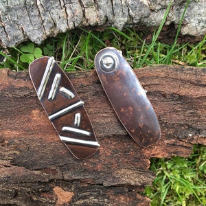 May include: Two copper and silver earrings with geometric designs. The earrings are oval shaped and have a hammered texture. One earring has a silver design of lines and rectangles. The other earring has a silver circle in the center.