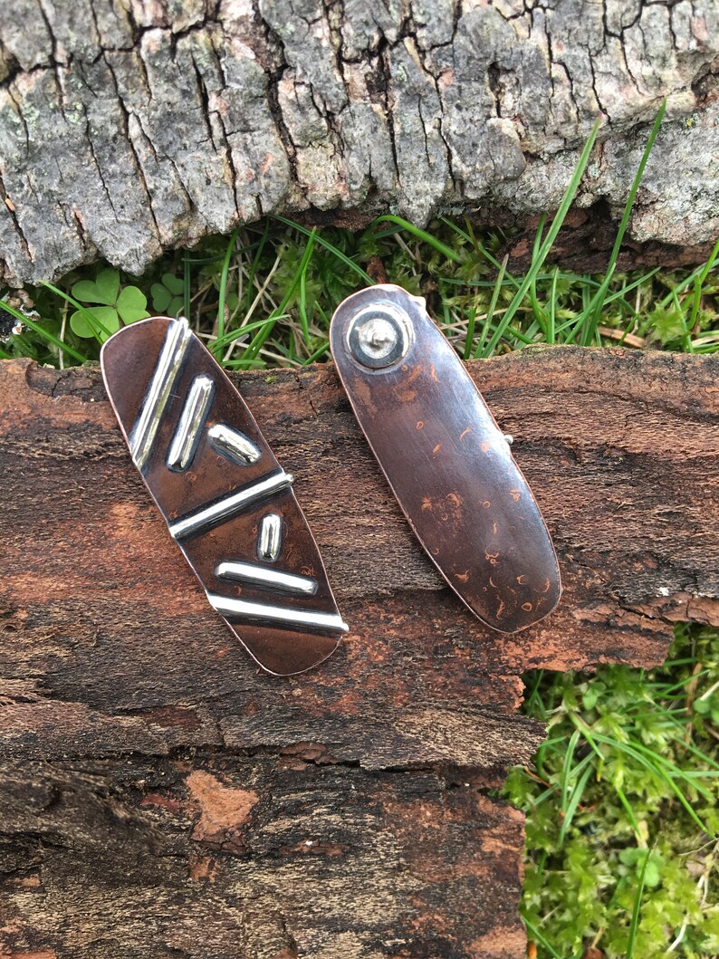 May include: Two copper and silver earrings with geometric designs. The earrings are oval shaped and have a hammered texture. One earring has a silver design of lines and rectangles. The other earring has a silver circle in the center.
