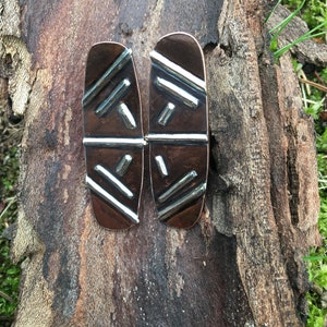 May include: A pair of copper earrings with a geometric pattern in silver. The earrings are oval shaped and have a textured surface.