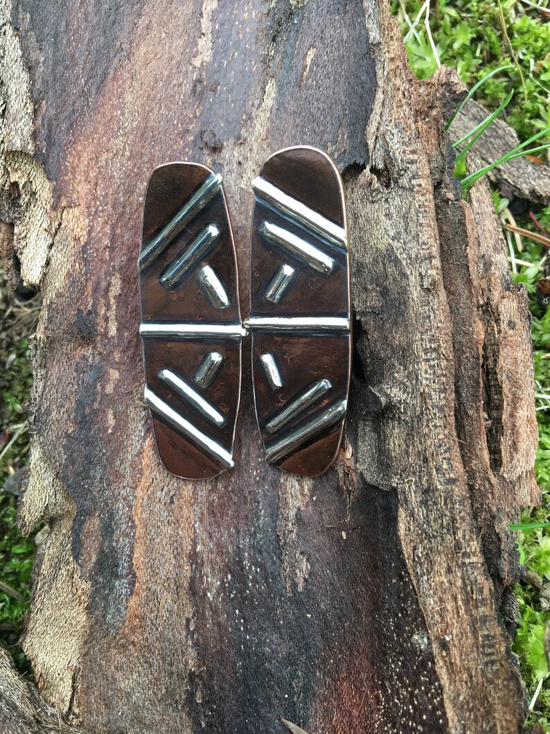 May include: A pair of copper earrings with a geometric pattern in silver. The earrings are oval shaped and have a textured surface.