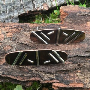May include: A pair of earrings with a geometric design. The earrings are made of copper and silver and have a dark brown color. The earrings are oval shaped and have a textured surface.