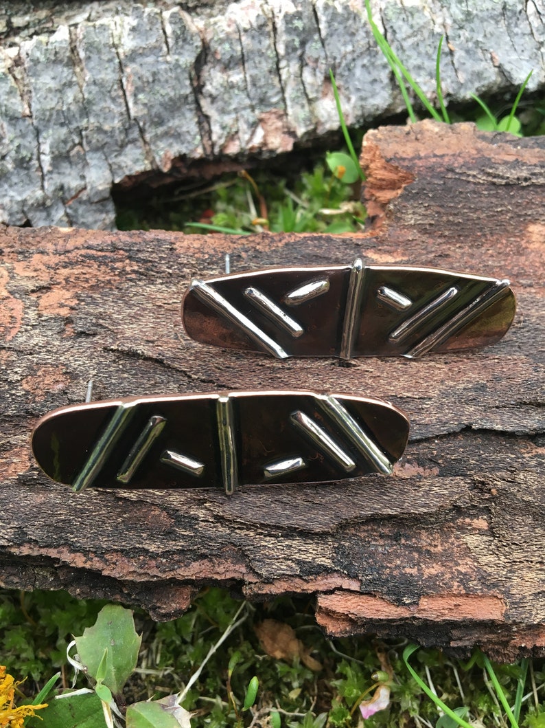 May include: A pair of earrings with a geometric design. The earrings are made of copper and silver and have a dark brown color. The earrings are oval shaped and have a textured surface.