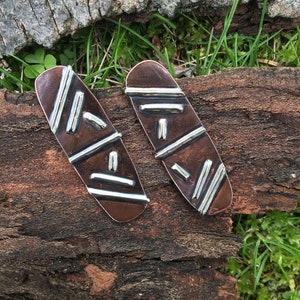 May include: A pair of copper and silver earrings with a geometric design. The earrings are oval shaped and have a hammered texture.