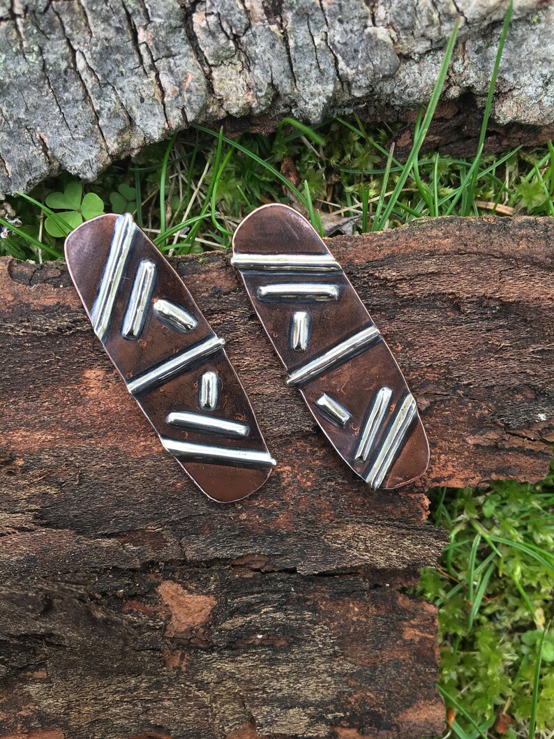 May include: A pair of copper and silver earrings with a geometric design. The earrings are oval shaped and have a hammered texture.