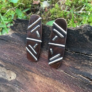 May include: A pair of silver and copper earrings with a geometric pattern. The earrings are oval shaped and have a textured surface.