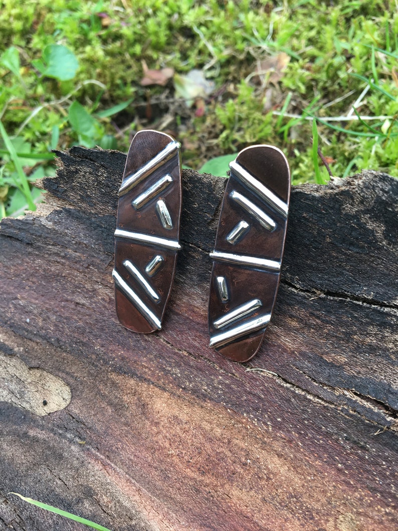 May include: A pair of silver and copper earrings with a geometric pattern. The earrings are oval shaped and have a textured surface.