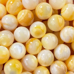 Pode incluir: Um punhado de contas redondas amarelas e brancas. As contas têm uma aparência marmorizada com variações de cor, que vão do amarelo pálido a um laranja mais intenso. As contas são provavelmente para fazer joias.
