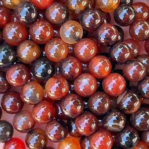 May include: A collection of round, polished beads in varying shades of deep red, burnt orange, and dark brown. The beads have a smooth, glossy finish with translucent qualities, showing natural internal patterns and color variations throughout.