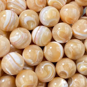 May include: A handful of round, polished beads in shades of tan and cream. The beads have a glossy finish and a swirling, marbled pattern. The beads are likely used for jewelry making or crafts.