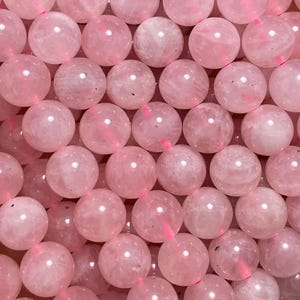 May include: A handful of light pink rose quartz beads. The beads are round and have a smooth, polished surface. The beads are strung together on a pink string. The background is blurred, with green leaves and white pebbles visible.