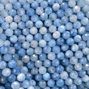 May include: A strand of small, round, faceted blue gemstone beads. The beads exhibit a range of light to medium blue tones with a semi-translucent quality and a reflective, cut surface texture, arranged in a dense, multi-row formation.