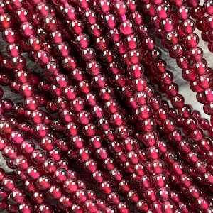 Könnte beinhalten: Nahaufnahme von mehreren Strängen kleiner, runder, tiefroter Perlen. Die Perlen sind aufgefädelt und erzeugen einen strukturierten und geschichteten Effekt. Die Perlen haben eine glänzende Oberfläche und reflektieren das Licht. Der Hintergrund ist eine neutrale graue Oberfläche.