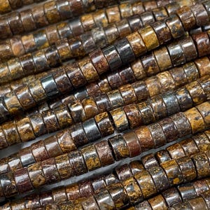 May include: Close-up of multiple strands of brown and gold gemstone beads. The beads are cylindrical with a slightly flattened shape, displaying a mix of earthy tones. The beads are arranged diagonally, creating a textured and detailed pattern.