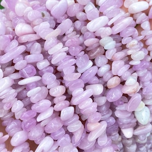 Puede incluir: Un puñado de cuentas de piedras preciosas de color púrpura claro. Las cuentas son pequeñas, de formas irregulares y están ensartadas. El color varía del lavanda pálido a un violeta más intenso. El fondo es follaje verde borroso.