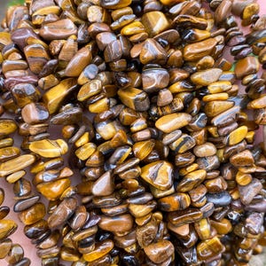 Pode incluir: Um punhado de lascas de pedras preciosas olho de tigre polidas em tons de castanho e dourado. As lascas são amarradas, criando uma estética natural e terrosa. As pedras preciosas são exibidas em um fundo verde desfocado.