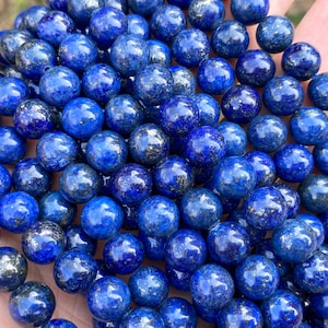 May include: A strand of deep blue lapis lazuli gemstone beads. The beads are round, polished, and have a speckled appearance. The beads are held in a hand, showcasing their color and texture. Ideal for jewelry making.