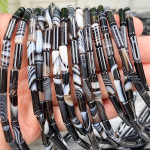 Puede incluir: Cadenas de cuentas de ágata bandeada en blanco y negro. Las cuentas cilíndricas están ensartadas, mostrando diversos patrones. La paleta de colores es principalmente negro, blanco y gris, con algunas cuentas translúcidas.
