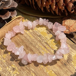 May include: A rose quartz chip bead bracelet. The bracelet is made of small, irregular, light pink stones strung together. The bracelet is resting on a weathered, light brown wooden surface, with pine cones and leaves in the background.