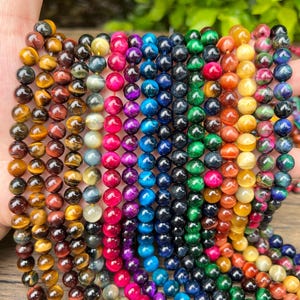 May include: A hand displays strands of colorful gemstone beads. The beads are round and come in a variety of colors, including brown, yellow, red, pink, blue, green, and purple. The beads are strung together, creating a vibrant and eye-catching display.