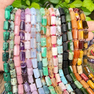 Puede incluir: Una mano muestra múltiples hilos de cuentas de piedras preciosas en varios colores. Las cuentas son rectangulares y están ensartadas, mostrando una diversa paleta de verdes, rosas, morados, azules, naranjas y marrones. Las cuentas son de diferentes tamaños y texturas.