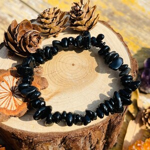 May include: A black beaded bracelet made of polished, irregular-shaped stones. The bracelet is displayed on a wooden surface with pine cones and other natural elements in the background. The stones are approximately 0.5 inches in length.