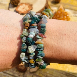 May include: A stack of three bracelets made of small, irregularly shaped, polished gemstones. The stones are primarily green, with accents of brown, white, and clear. The bracelets are worn on a wrist, with a blurred background.