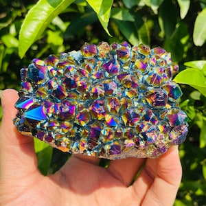 Puede incluir: Un cúmulo de cristales de amatista iridiscentes con un arcoíris de colores, incluyendo morado, azul, verde y dorado. Los cristales están agrupados en una formación natural.