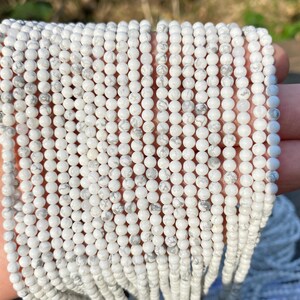 May include: Strands of small, round, white beads with gray marbling. The beads are strung together, creating a delicate and textured appearance. The beads are held in a hand, suggesting they are ready for use in jewelry making or crafts.