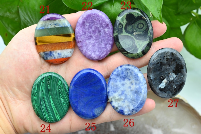 May include: Seven oval-shaped polished stones, each with a different color and pattern. The stones are arranged in a row on a hand, with the numbers 21 through 27 written below each stone.