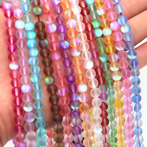 Puede incluir: Surtido de cuentas de vidrio esmerilado iridiscente en varios colores, incluyendo rosa, azul, morado, verde y marrón. Las cuentas son redondas y tienen un acabado liso y mate.