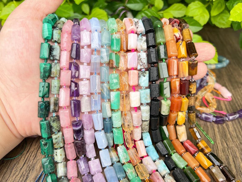 May include: A hand displays multiple strands of gemstone beads in various colors. The beads are rectangular and strung together, showcasing a diverse palette of greens, pinks, purples, blues, oranges, and browns. The beads are of different sizes and textures.