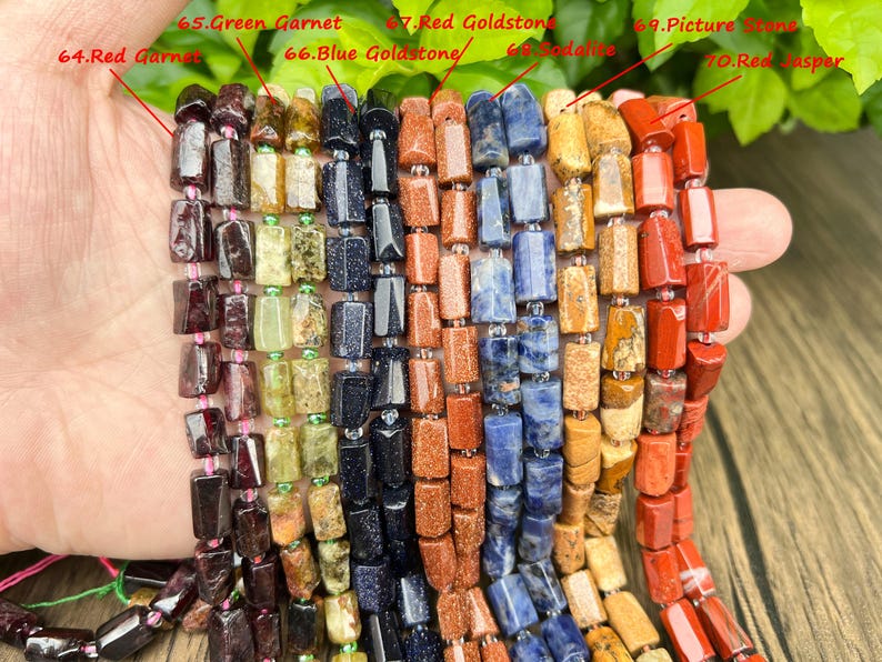 May include: Close-up of multiple strands of gemstone beads in various colors. The beads include red garnet, green garnet, blue goldstone, sodalite, picture stone, and red jasper. Each bead is rectangular and strung together.