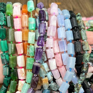 May include: A hand holding strands of colorful gemstone beads. The beads are rectangular and come in various colors, including green, pink, purple, blue, and black. The beads are strung together with string, and the background is a light brown.