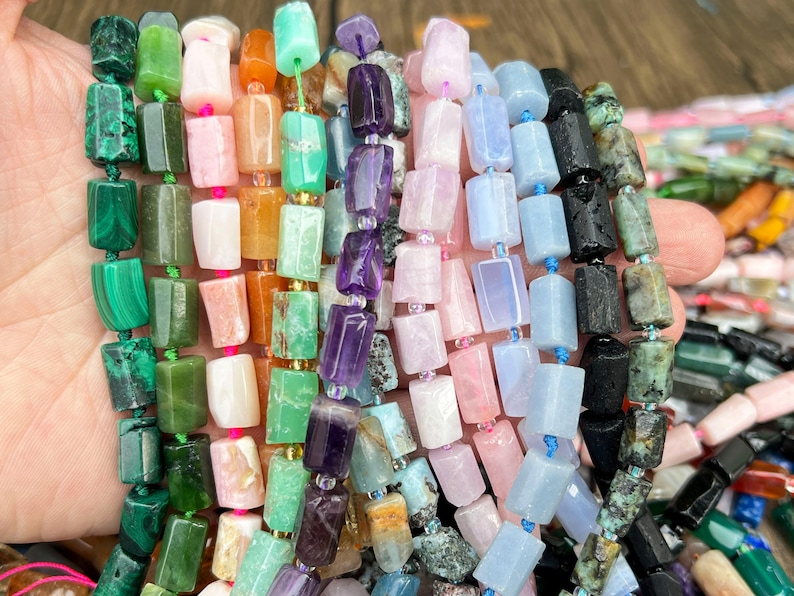 May include: A hand holding strands of colorful gemstone beads. The beads are rectangular and come in various colors, including green, pink, purple, blue, and black. The beads are strung together with string, and the background is a light brown.