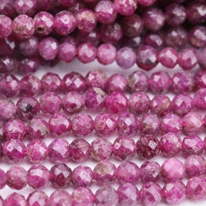 May include: Close-up of multiple strands of faceted, round, deep pink ruby beads. The beads are strung together, showcasing their polished surfaces and variations in color, ranging from light to dark pink.