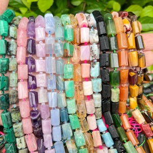 May include: A collection of colorful gemstone beads in various shades of green, pink, purple, blue, orange, and black. The beads are rectangular and strung together, showcasing a variety of natural stone textures and colors. The beads are held in a hand.