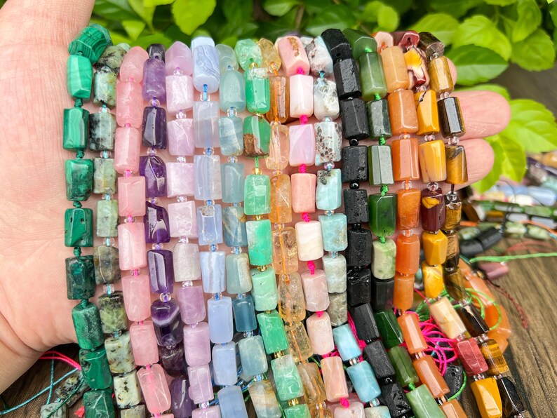 May include: A collection of colorful gemstone beads in various shades of green, pink, purple, blue, orange, and black. The beads are rectangular and strung together, showcasing a variety of natural stone textures and colors. The beads are held in a hand.