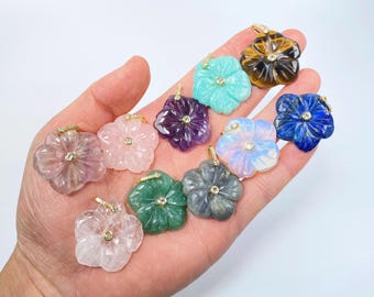 May include: A collection of flower-shaped gemstone pendants in various colors, including pink, green, blue, and purple. Each pendant has a gold bail and a small, sparkling accent in the center. The pendants are displayed on a hand.