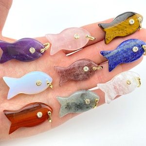 May include: A collection of gemstone fish pendants in various colors, including purple, pink, blue, and brown. Each fish has a gold-colored bail and a small, round, clear stone. The pendants are displayed on a light background.