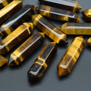 May include: A collection of polished tiger's eye crystal wands. The wands are a mix of golden yellow and brown, with a glossy finish. Each wand has a hexagonal shape and pointed ends, perfect for crystal healing and meditation.
