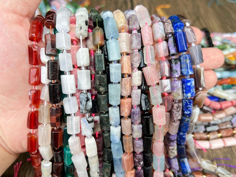 May include: Strands of colorful gemstone beads in various shapes and sizes. The beads are a mix of colors, including red, white, black, green, blue, and purple. The beads are strung together with a clear string.