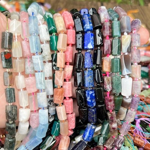 May include: Strands of colorful gemstone beads in various shapes and sizes. The beads are in shades of pink, blue, green, black, and white. The beads are strung together with pink and blue string. The beads are held in a hand.