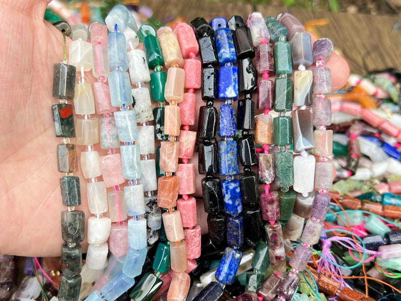 May include: Strands of colorful gemstone beads in various shapes and sizes. The beads are in shades of pink, blue, green, black, and white. The beads are strung together with pink and blue string. The beads are held in a hand.