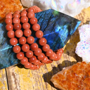 Puede incluir: Primer plano de un brazalete de cuentas de varias hebras hecho de cuentas de piedra arenisca rojo-marr&oacute;n natural. El brazalete est&aacute; sobre un cristal de labradorita azul, rodeado de otros cristales, incluyendo un c&uacute;mulo de cuarzo blanco y un c&uacute;mulo de citrino amarillo.